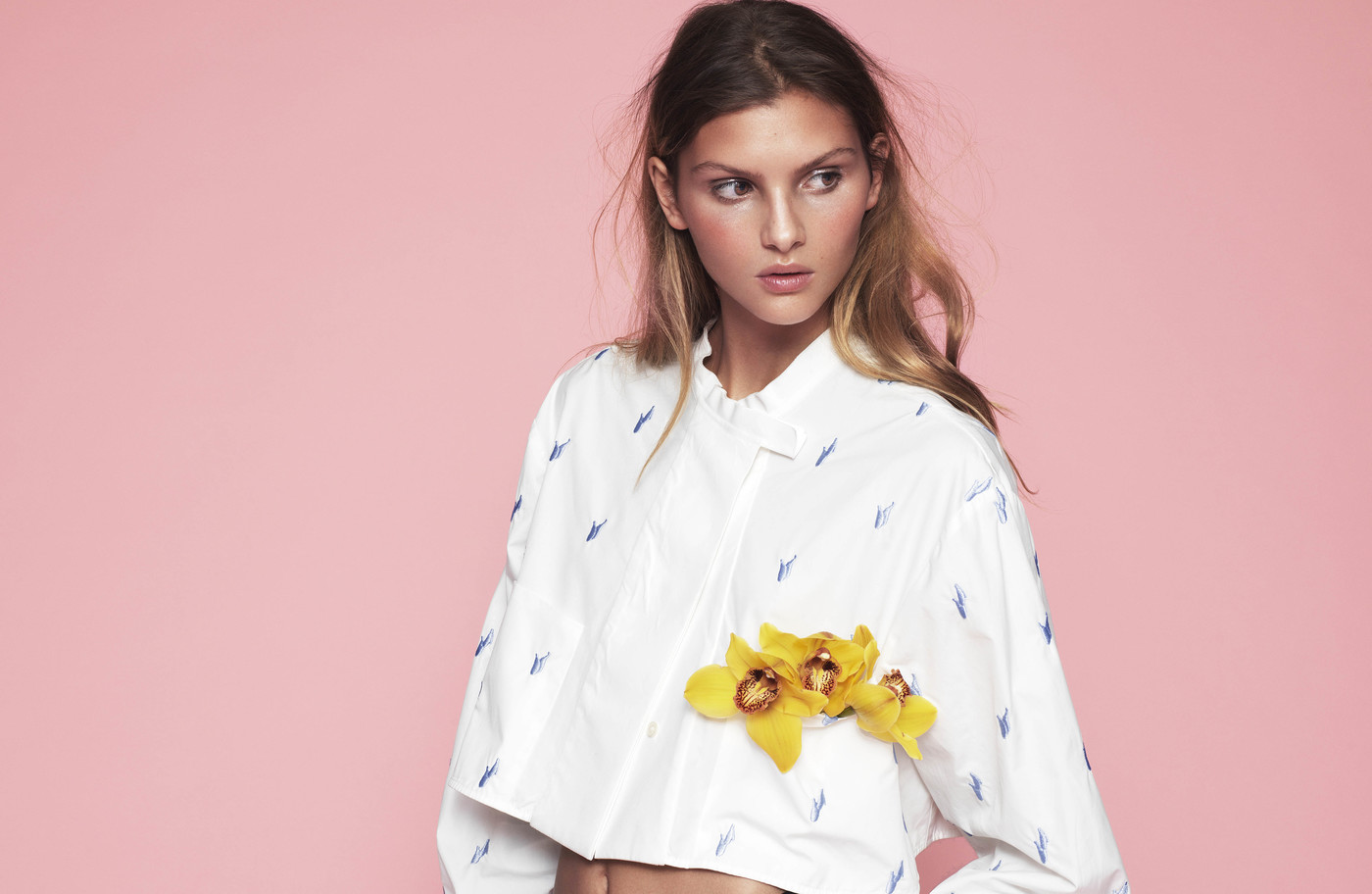 Young woman in a short patterned white button-down shirt posing in front of a pink background, decorated with yellow flowers.