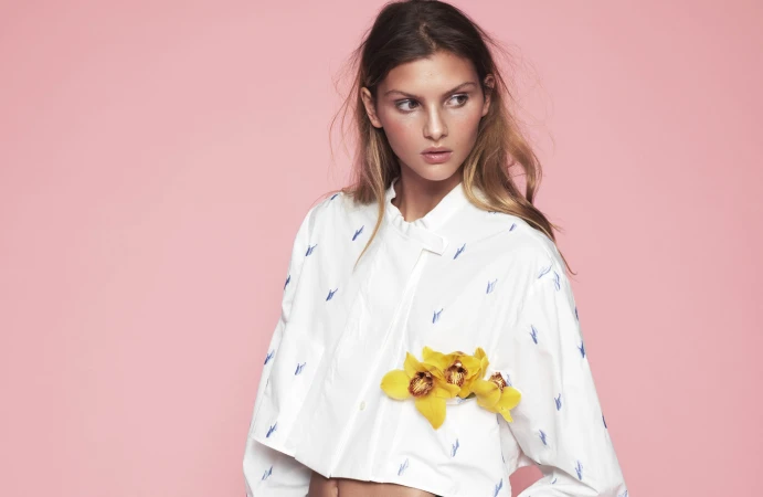 Young woman in a short patterned white button-down shirt posing in front of a pink background, decorated with yellow flowers.