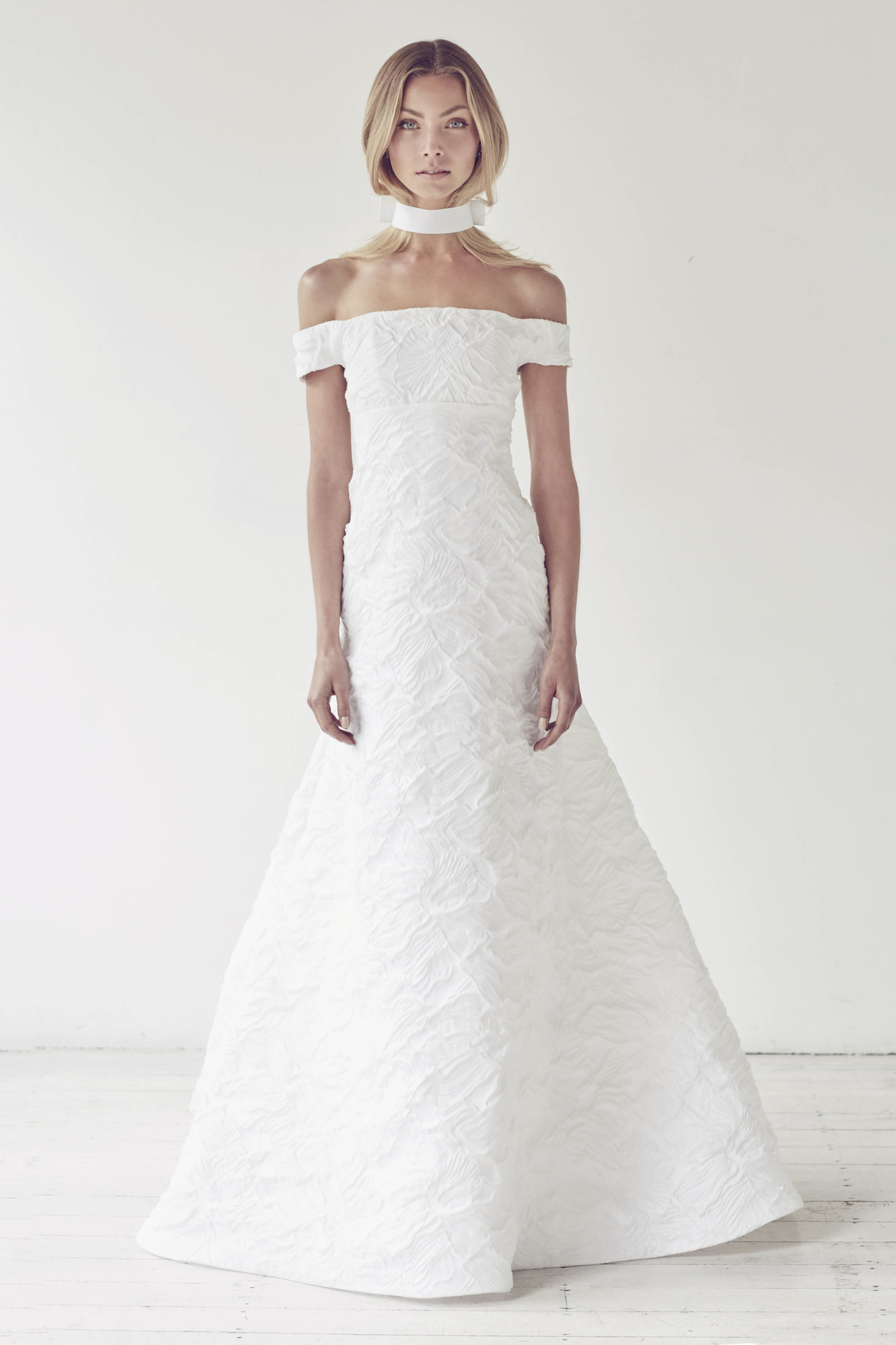 A woman models a white, off-the-shoulder wedding dress in a studio. She wears a white choker necklace and has blonde hair.