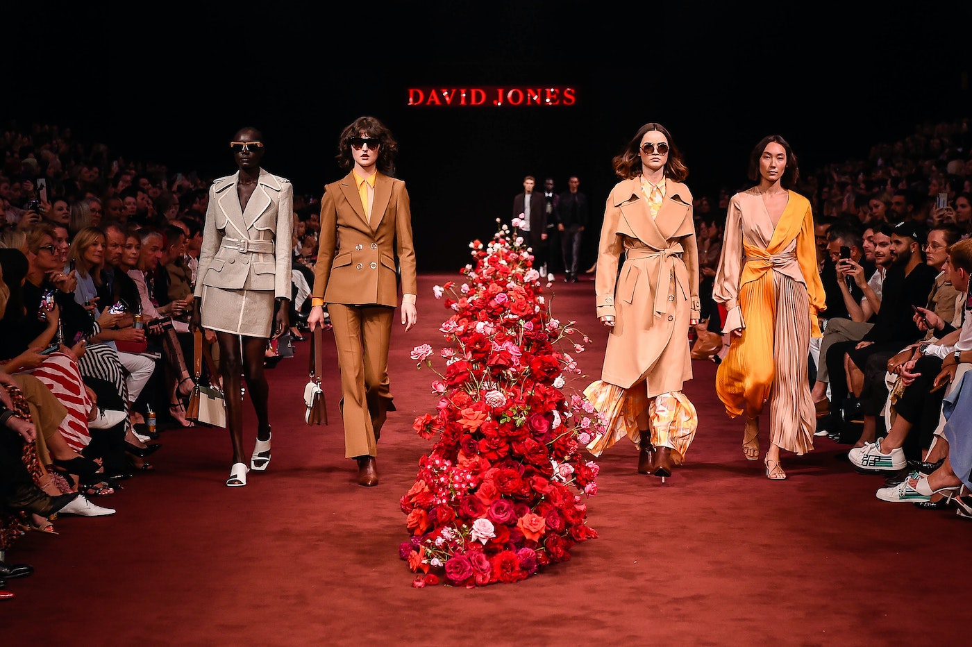 Fashion show on a red runway with four models wearing suits and dresses, a floral arrangement, and a crowd in the background.