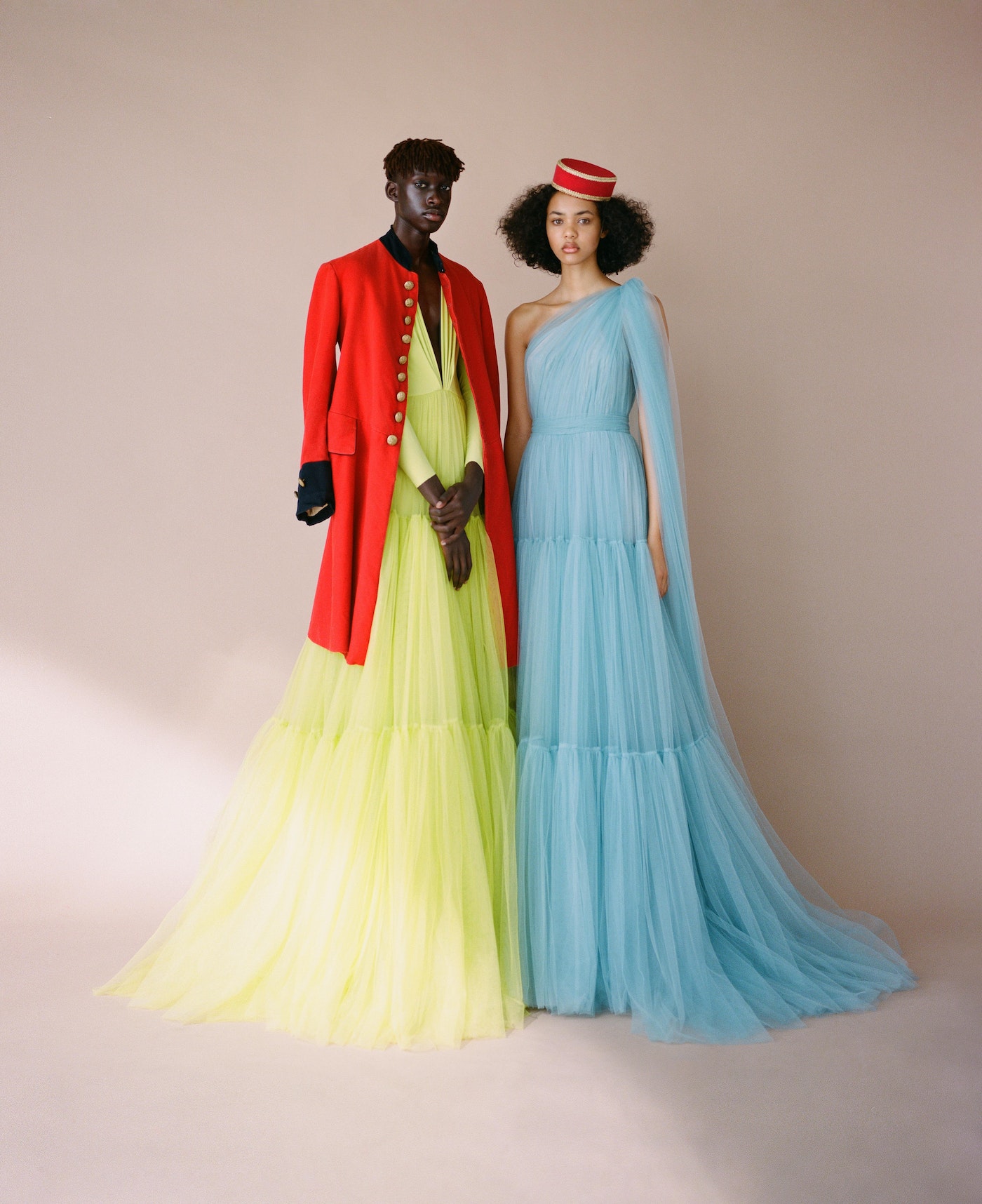Two fashion models wearing gowns in front of a neutral backdrop. One model wears a red jacket and the other a red hat.