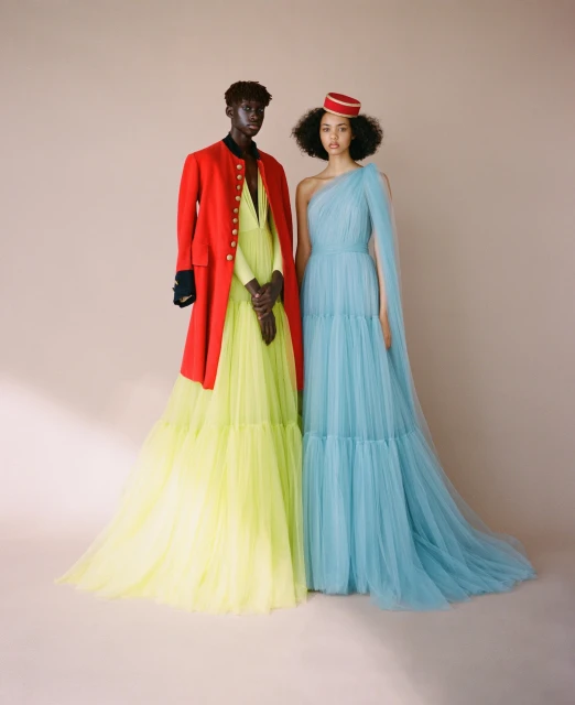 Two fashion models wearing gowns in front of a neutral backdrop. One model wears a red jacket and the other a red hat.