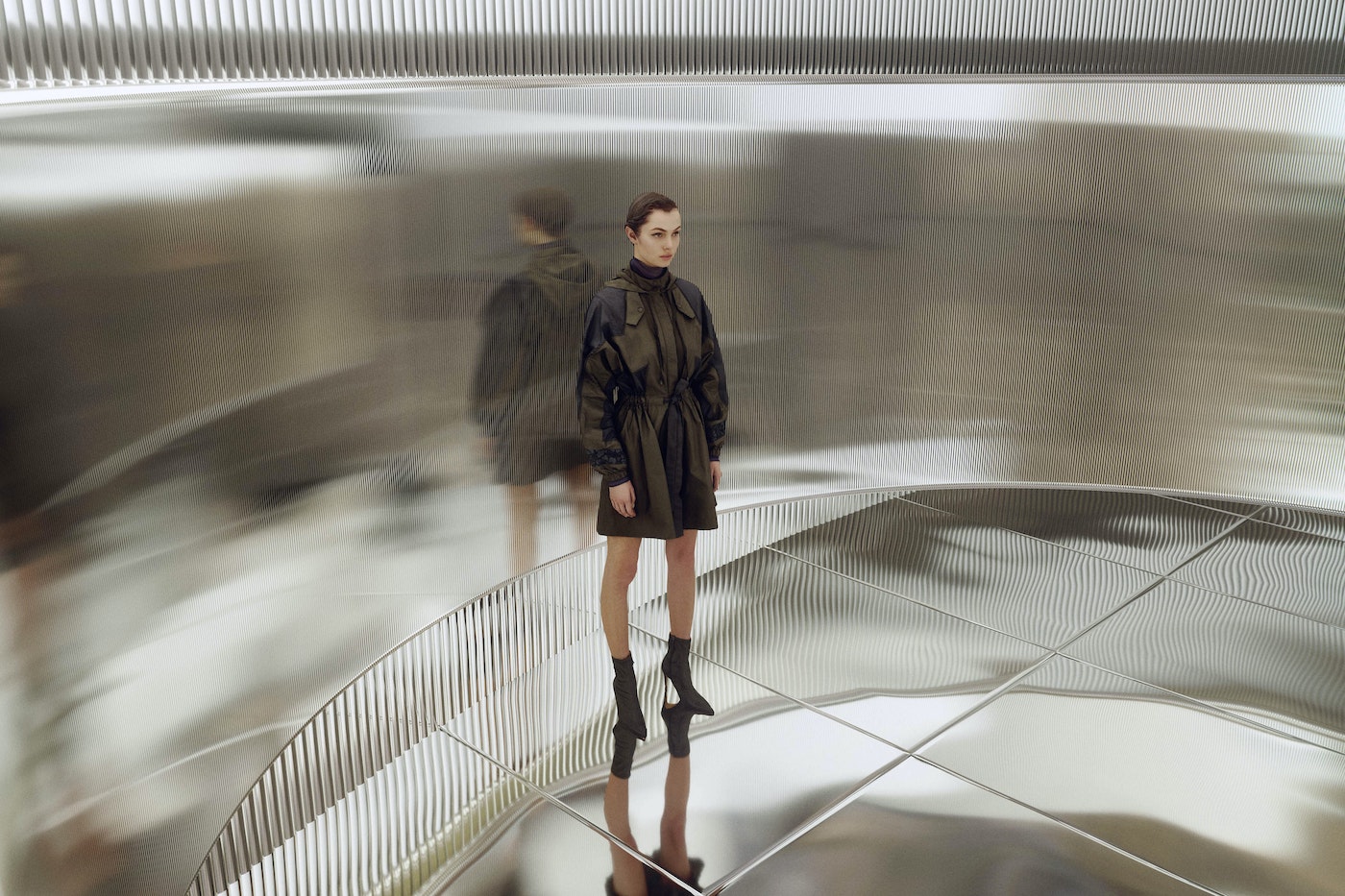 Fashionable adult woman poses in a dark green jacket dress, black heeled sock boots against a metallic abstract architectural backdrop.
