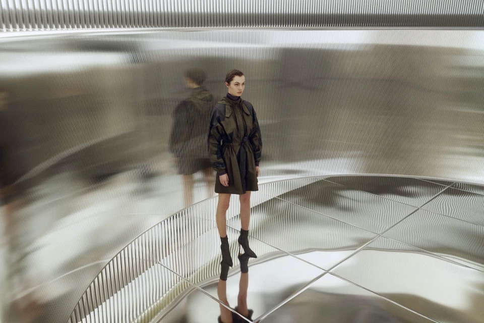Fashionable adult woman poses in a dark green jacket dress, black heeled sock boots against a metallic abstract architectural backdrop.