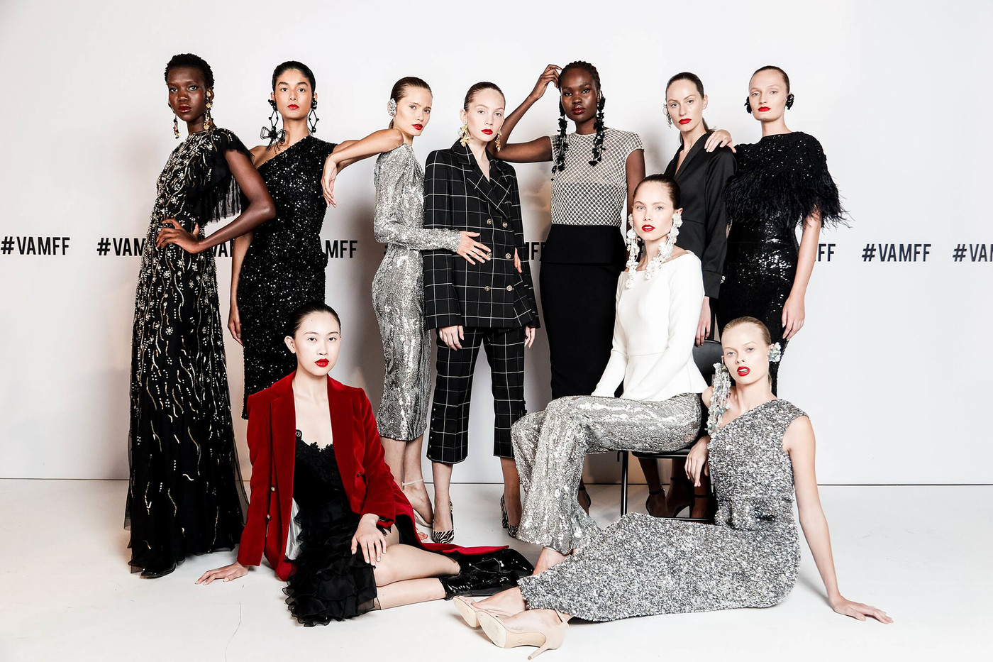 Diverse group of nine fashion models in dresses and pantsuits pose in a photo studio against a white backdrop; runway fashion show.