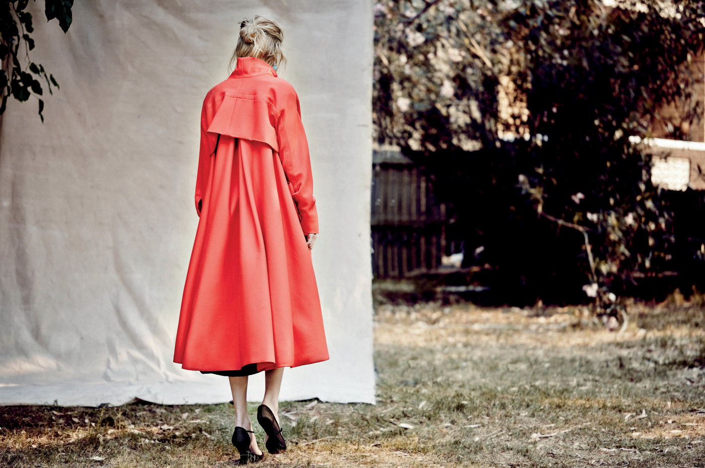 Back view of a blonde woman in a bright red coat walking on grass towards trees and a white backdrop, wearing black heels.