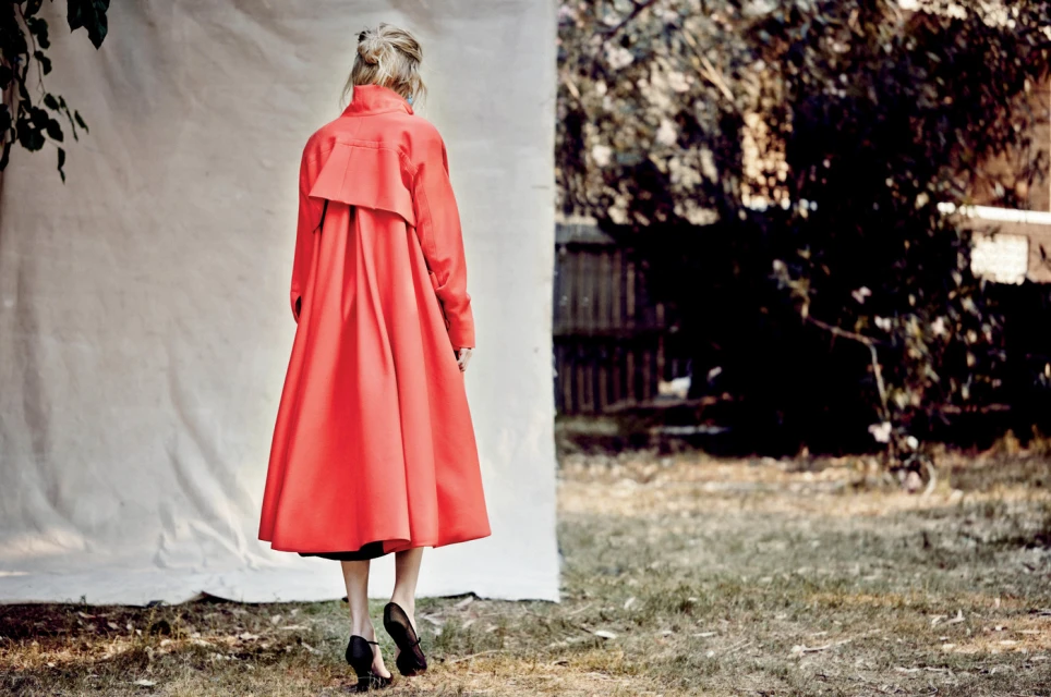 Back view of a blonde woman in a bright red coat walking on grass towards trees and a white backdrop, wearing black heels.