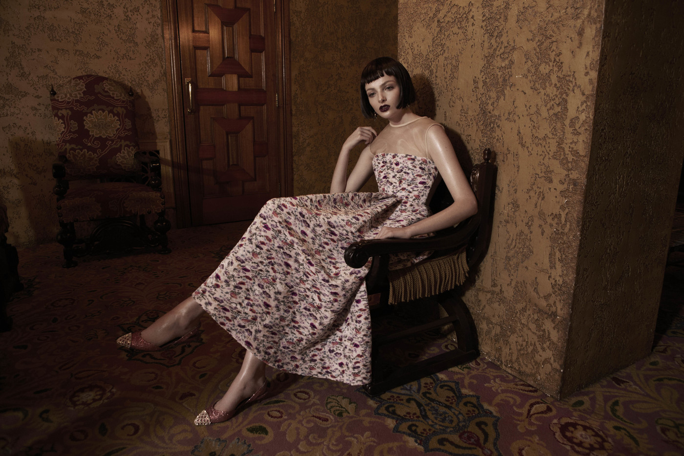 Elegant woman in floral dress poses in antique chair. The scene features textured walls and patterned carpet, creating a vintage atmosphere.