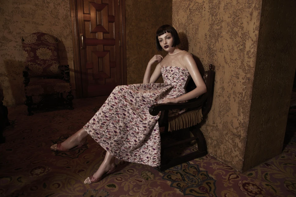 Elegant woman in floral dress poses in antique chair. The scene features textured walls and patterned carpet, creating a vintage atmosphere.