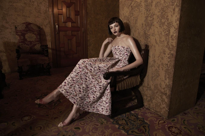 Elegant woman in floral dress poses in antique chair. The scene features textured walls and patterned carpet, creating a vintage atmosphere.