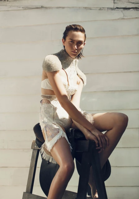 A woman in an ornate dress sits on a saddle stand against a rustic white wood wall in a sunlit setting, showcasing fashion and style.