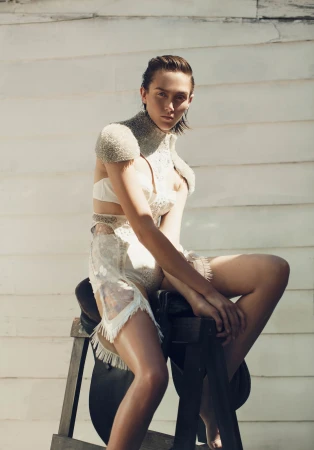 A woman in an ornate dress sits on a saddle stand against a rustic white wood wall in a sunlit setting, showcasing fashion and style.