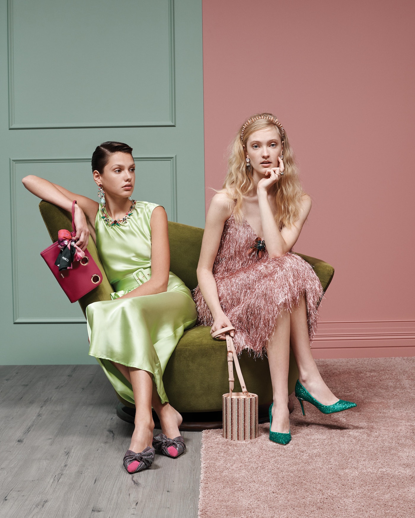 Two women seated in a vintage chair. One wears a green dress and the other wears a pink dress with fringe. Both hold designer handbags.