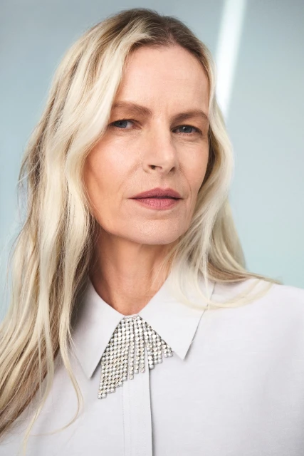 Close-up studio portrait of an adult woman with blonde hair wearing a light-grey collared shirt with a silver, diamond-like adornment.