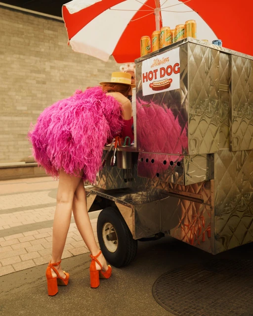 Woman in pink faux-feather dress and orange platform shoes at a hot dog stand with red-and-white umbrella in an outdoor setting.