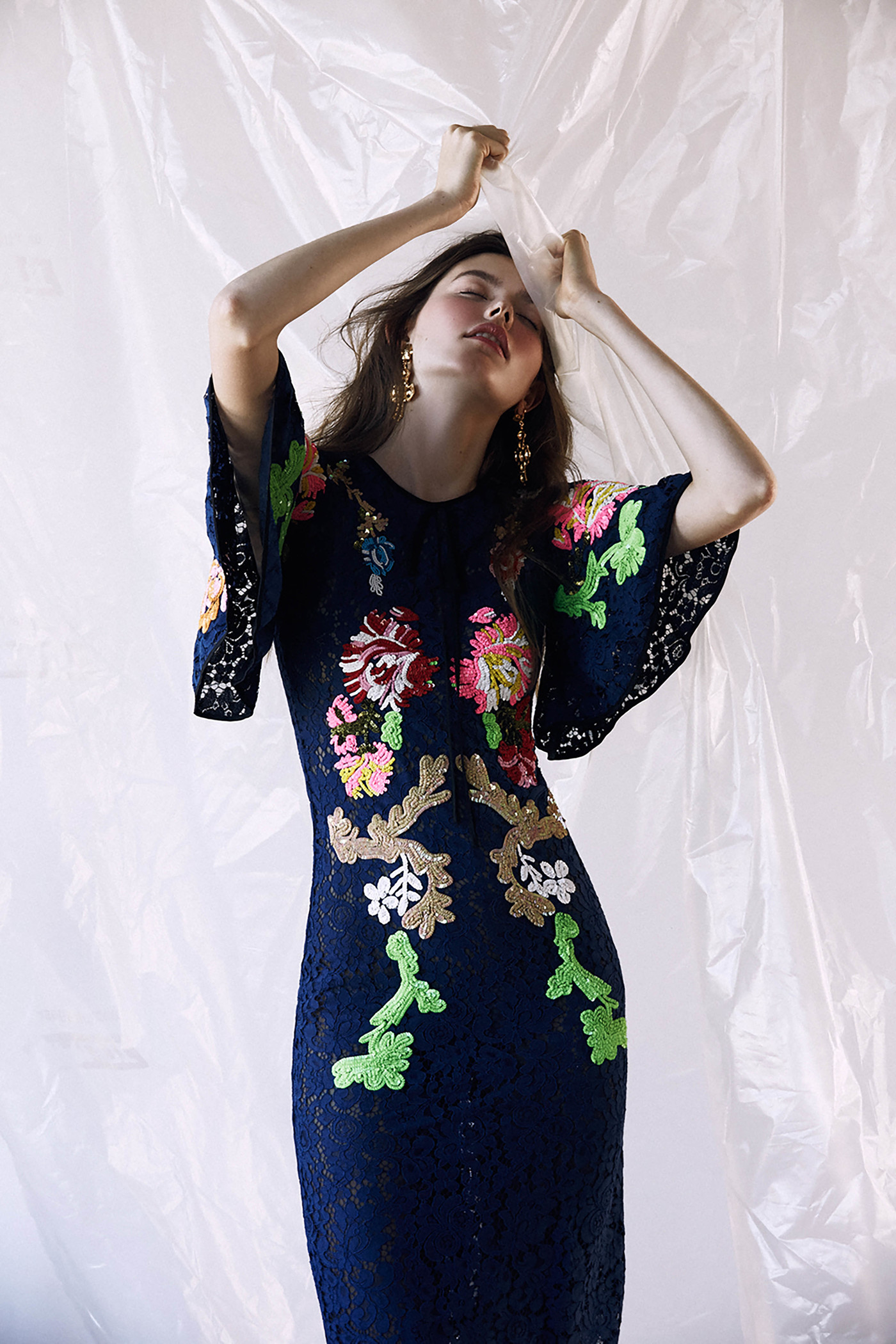 A woman poses in a fitted, knee-length navy dress with colorful floral embroidery, sheer sleeves, and gold earrings, against a white backdrop.