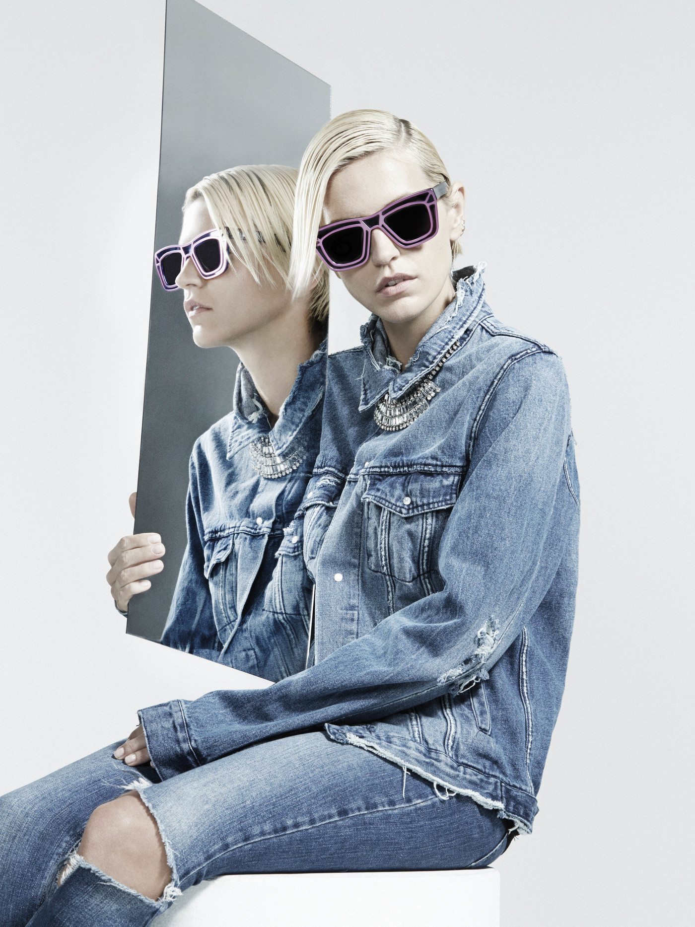 Model in denim jacket wearing statement sunglasses and holding a tall rectangular mirror reflecting her profile