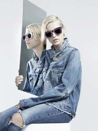 Model in denim jacket wearing statement sunglasses and holding a tall rectangular mirror reflecting her profile