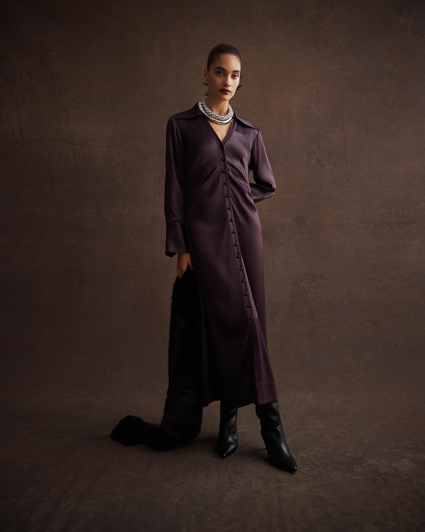 Elegant woman in a long-sleeved dark purple dress and black boots poses against a brown background, holding a fur garment.