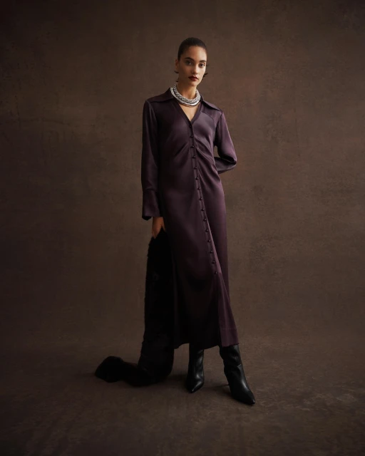 Elegant woman in a long-sleeved dark purple dress and black boots poses against a brown background, holding a fur garment.