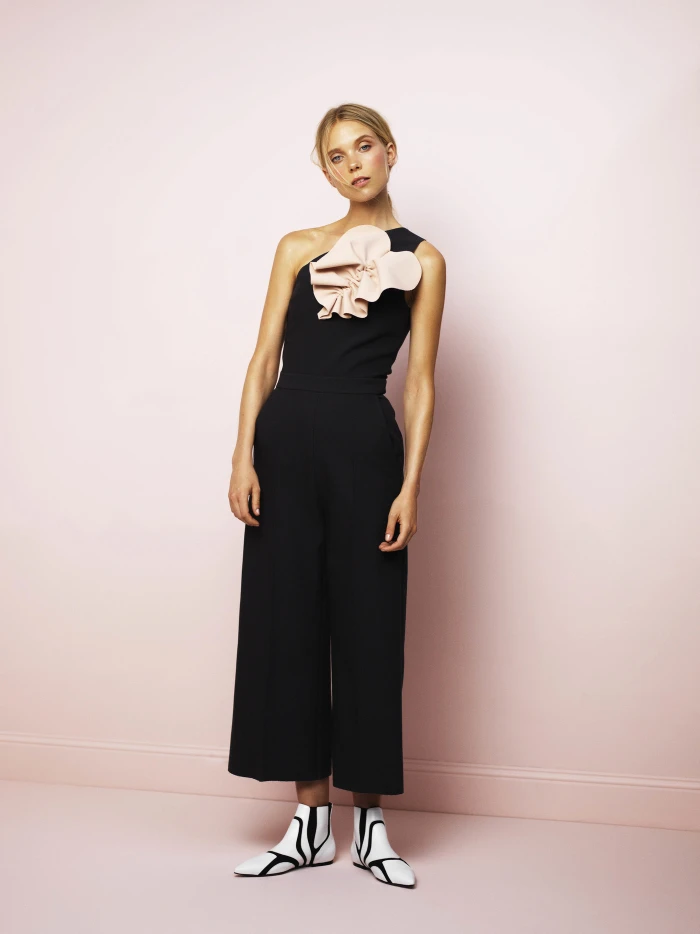 Model in black jumpsuit with pink floral appliqué and patterned boots standing against a pale pink backdrop