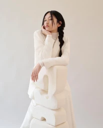 East Asian woman in white blazer resting chin on hand beside stacked white foam sculptures against a plain backdrop.