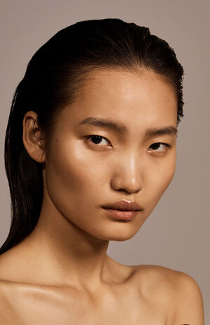 Close-up studio shot of a tanned Asian female model with wet dark hair, shiny skin and lips against a neutral grey background.