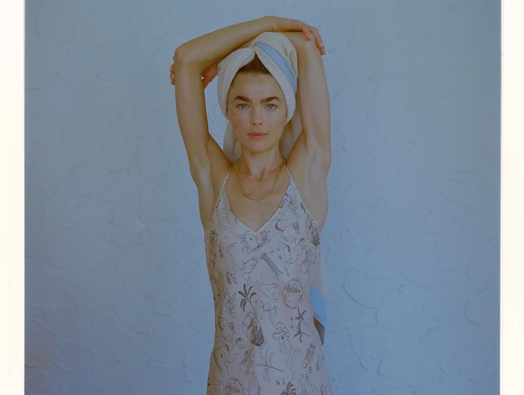 Woman in a slip dress and towel-wrapped hair strikes a pose against a textured blue wall in a naturally lit indoor setting.