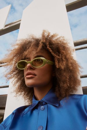 Close-up of a person with curly hair wearing stylish olive green sunglasses and a blue shirt against an abstract white backdrop.