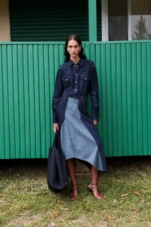 A woman in a denim shirt and patchwork denim skirt stands outside a green building with a black tote bag and strappy heeled sandals.