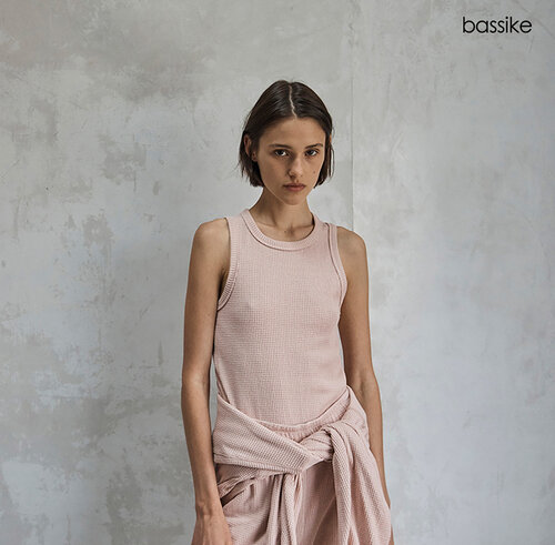 A slender woman poses indoors wearing a pink tank top and draped shirt against a textured gray wall, with Bassike logo in the corner.
