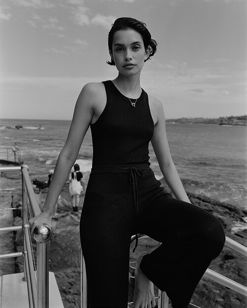 Monochrome portrait of a woman in a black tank top and pants, posing by a railing with ocean and sky in the background.