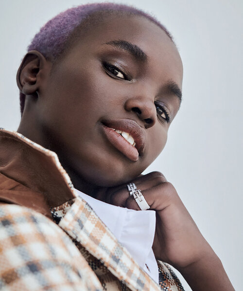 Close-up studio portrait of a young adult woman with short purple hair wearing a tan and brown plaid jacket looking at the camera.