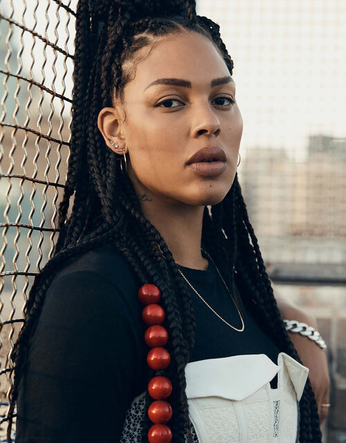 Portrait of a stylish adult woman with braided hair wearing a dark top and decorative white corset against an urban background.