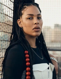 Close-up portrait of a woman with long braided hair wearing a dark top, white structured corset, and red bead necklace.