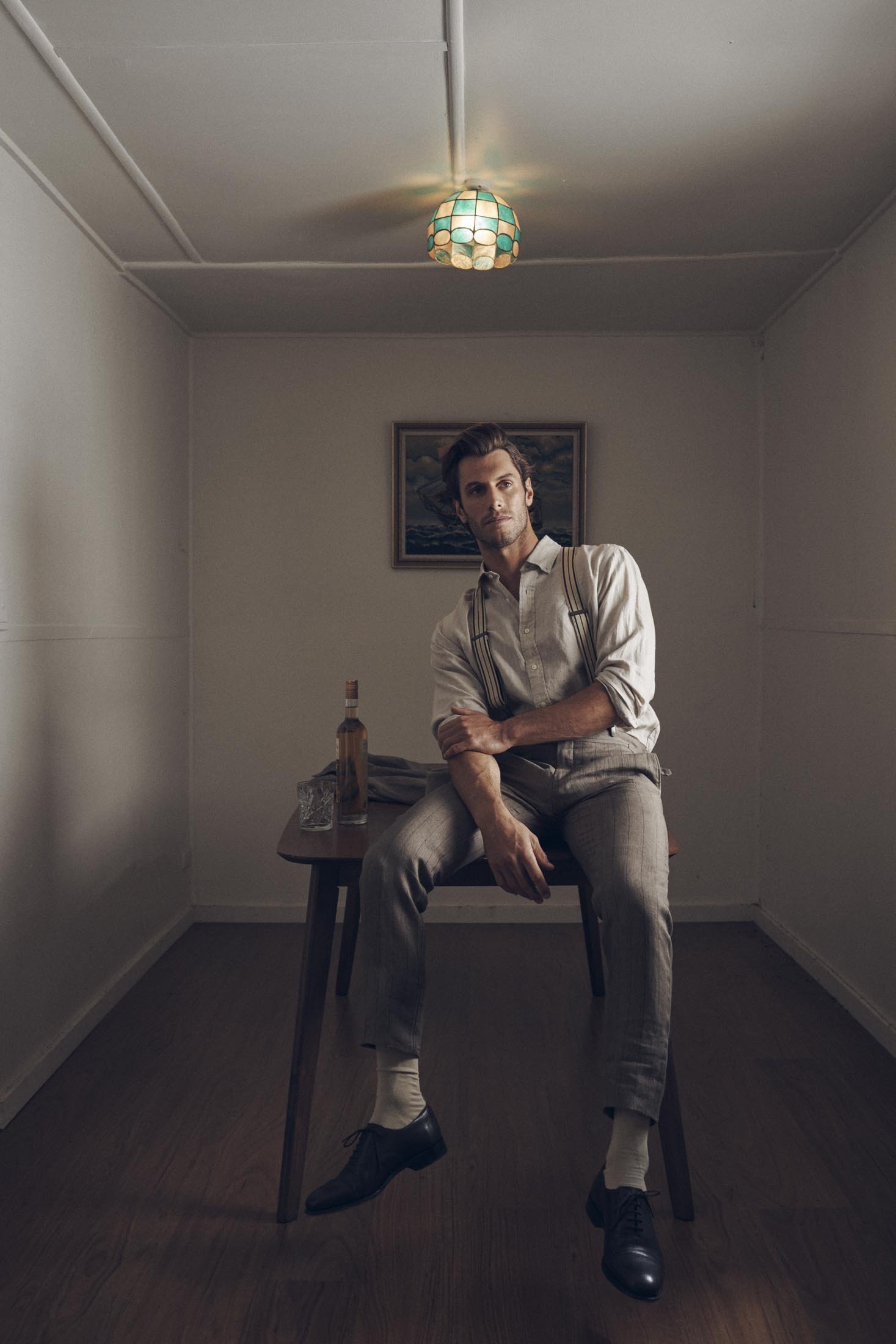 Man in button-down shirt and suspenders sitting on table in dimly lit room with bottle and glass. Interior scene with neutral colors.