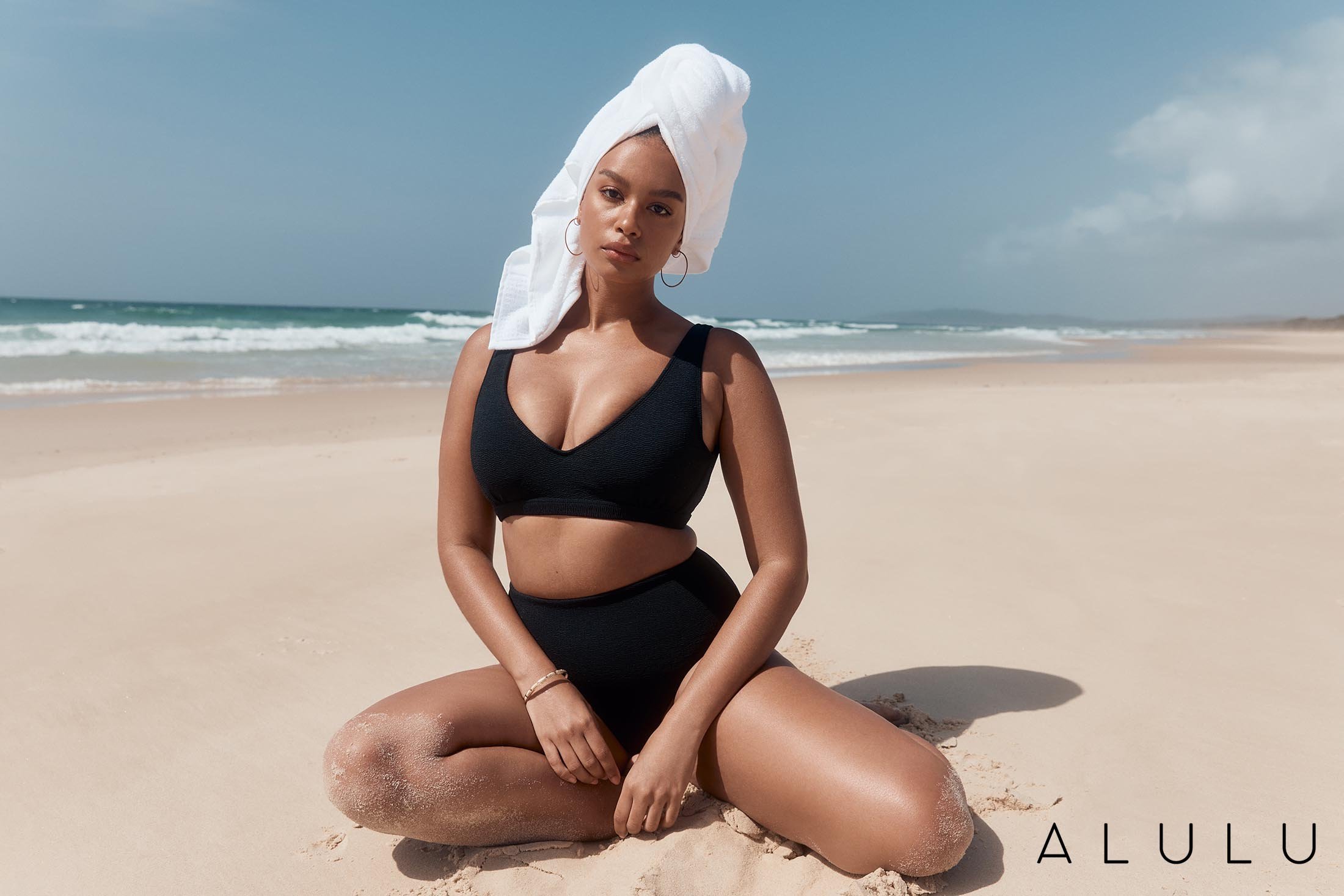 Woman in black bikini and towel on head sits on sandy beach near ocean on sunny day for fashion or beauty advertising.