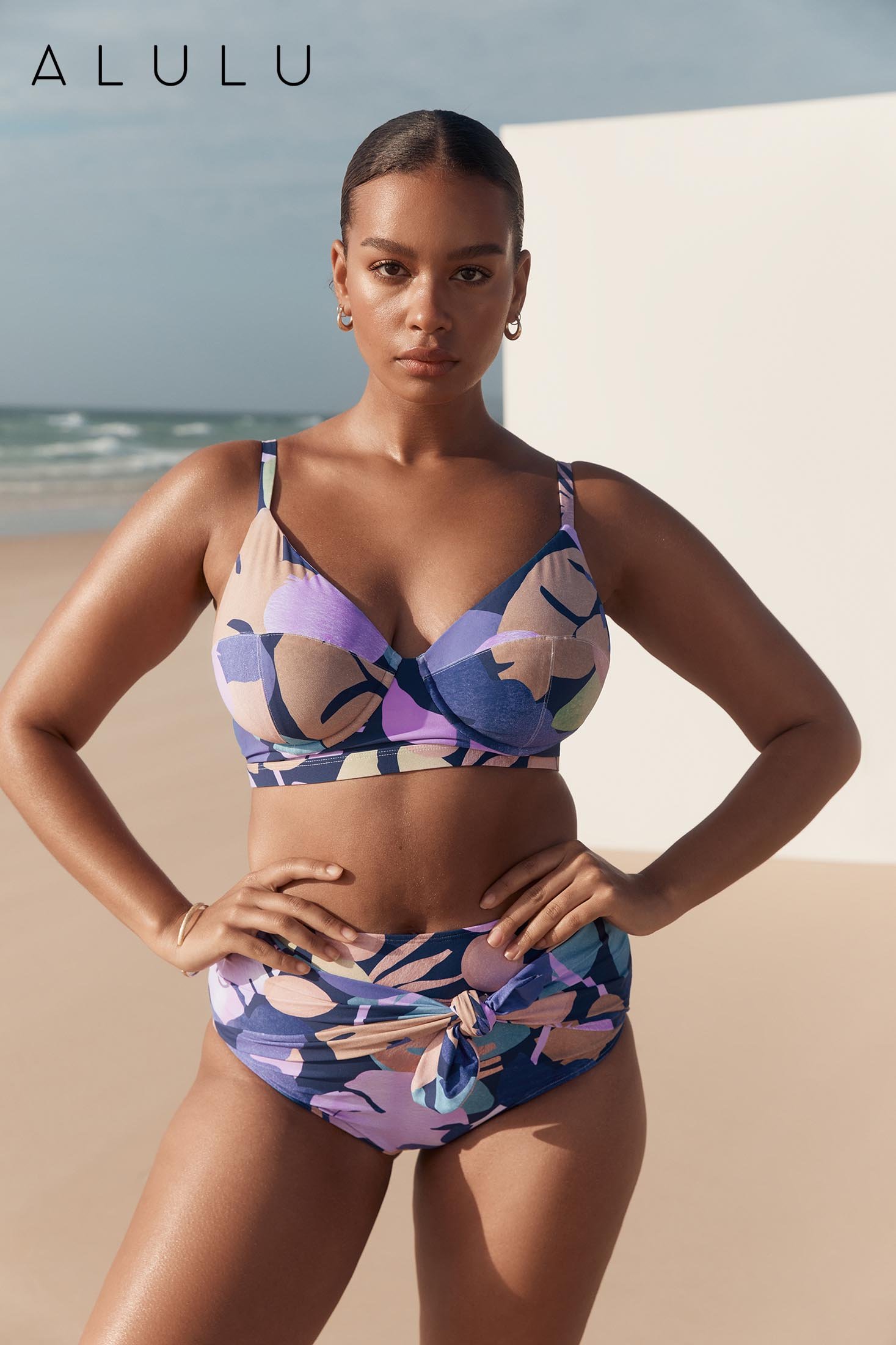 Woman in a floral bikini poses confidently on a beach with sand and water behind her in a promotional outdoor shot by Alulu.