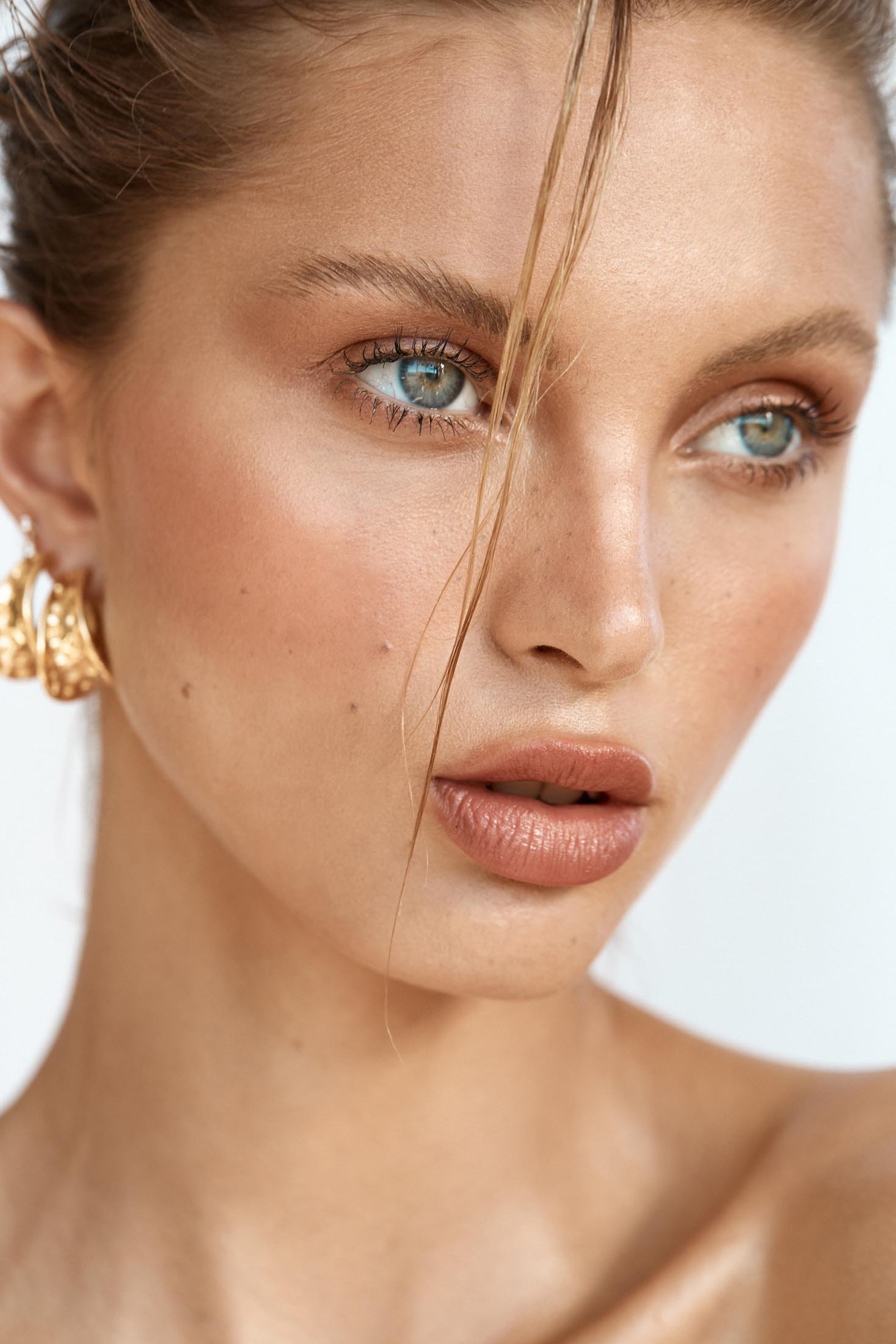 Close-up portrait of a beautiful fair-skinned woman with light eyes and highlighted makeup wearing gold earrings against a plain white backdrop.