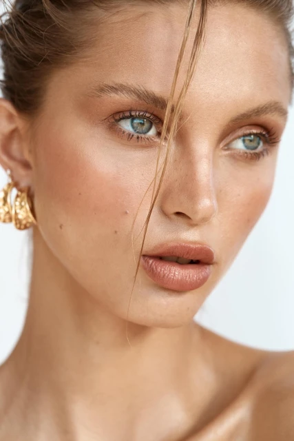 Close-up portrait of a beautiful fair-skinned woman with light eyes and highlighted makeup wearing gold earrings against a plain white backdrop.