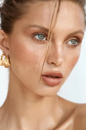 Close-up portrait of a beautiful fair-skinned woman with light eyes and highlighted makeup wearing gold earrings against a plain white backdrop.