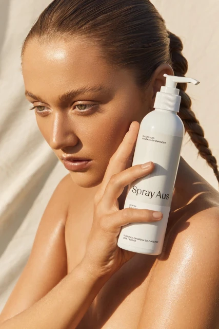 Close-up of a woman with a bare shoulder, fair skin and braided hair holding a white bottle of sunless tanning lotion.