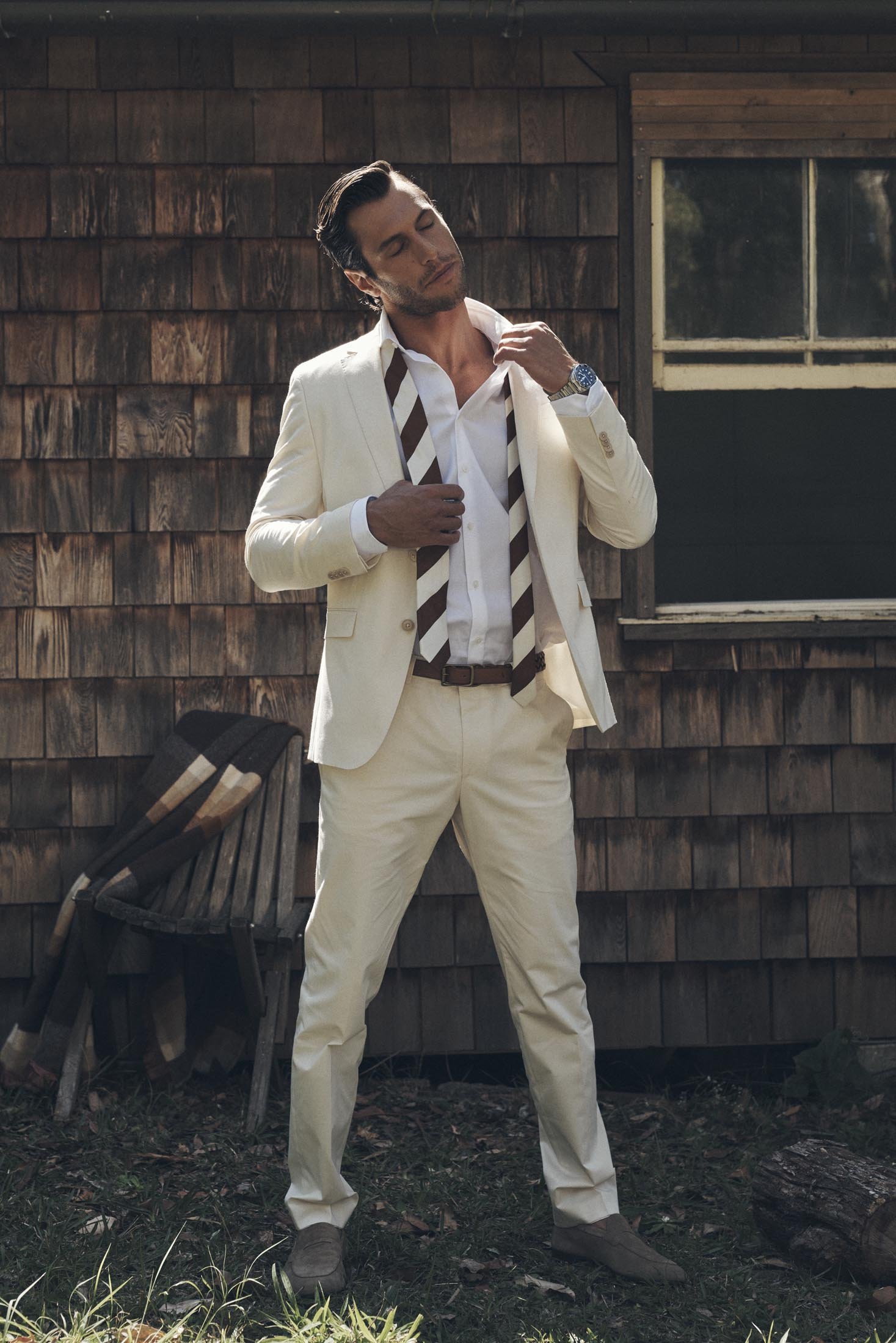 Man in a light suit stands in front of a wood-paneled wall with a window, accessorized with a striped tie and watch. Outdoor setting.
