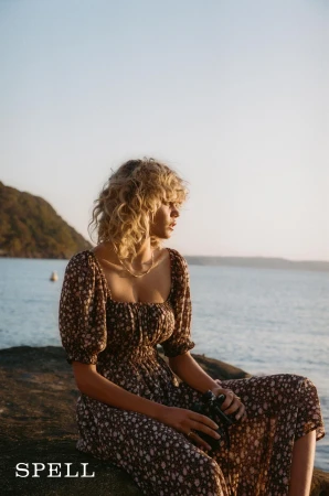 Woman in a floral dress sitting on a rock near the ocean at sunset; she has blonde curly hair and wears a gold necklace.  
