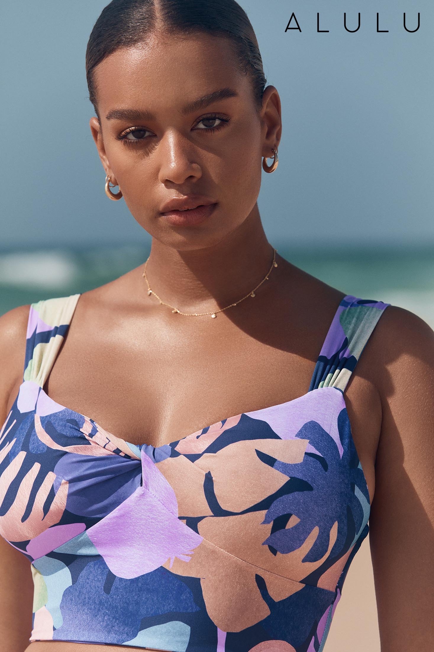 A woman in a tropical-print bikini top poses in front of an ocean backdrop, showcasing gold hoop earrings and a delicate necklace.