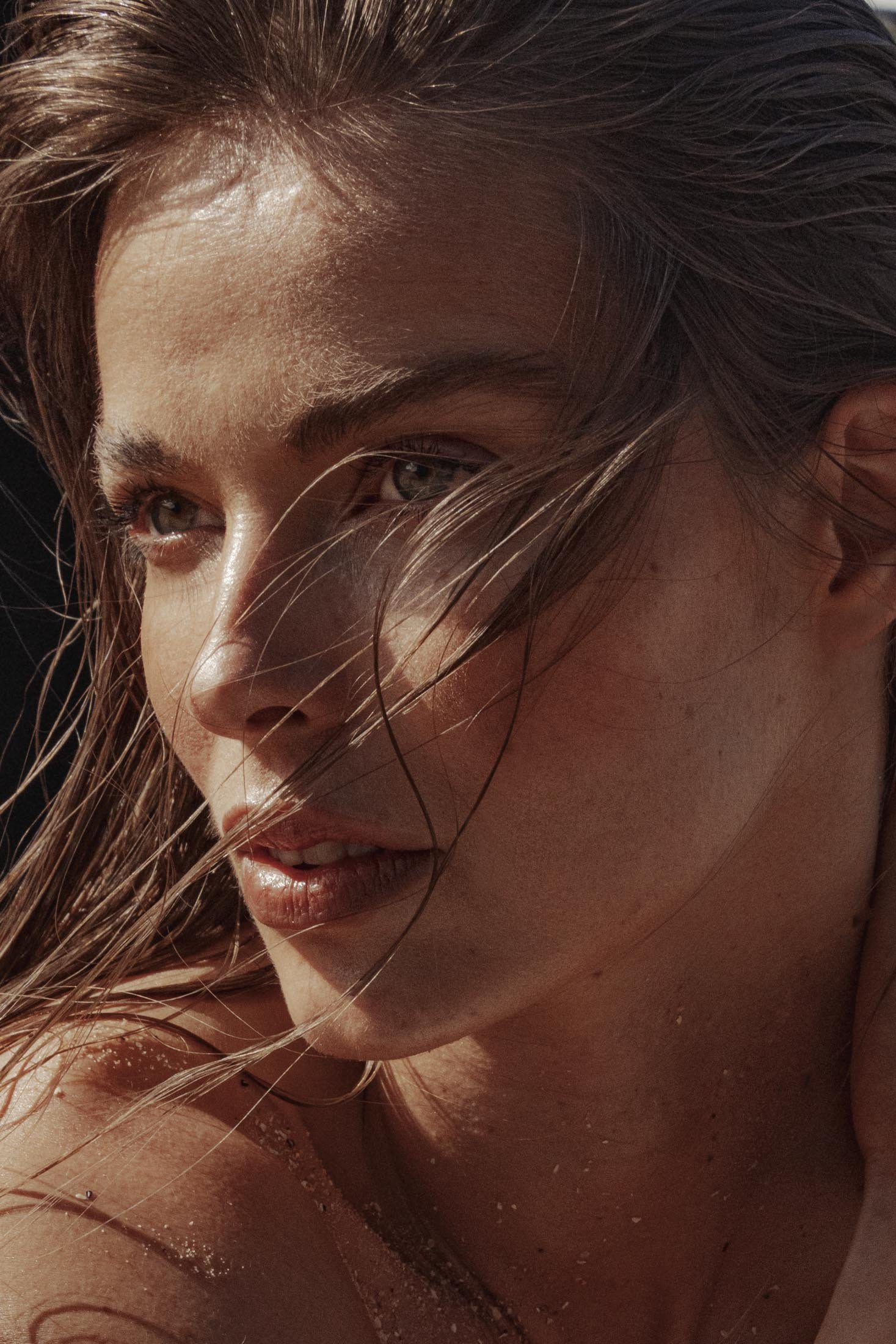 Close-up portrait of a young adult woman with sun-kissed skin, wet hair, and subtle makeup gazing off-frame, bathed in warm, natural light.