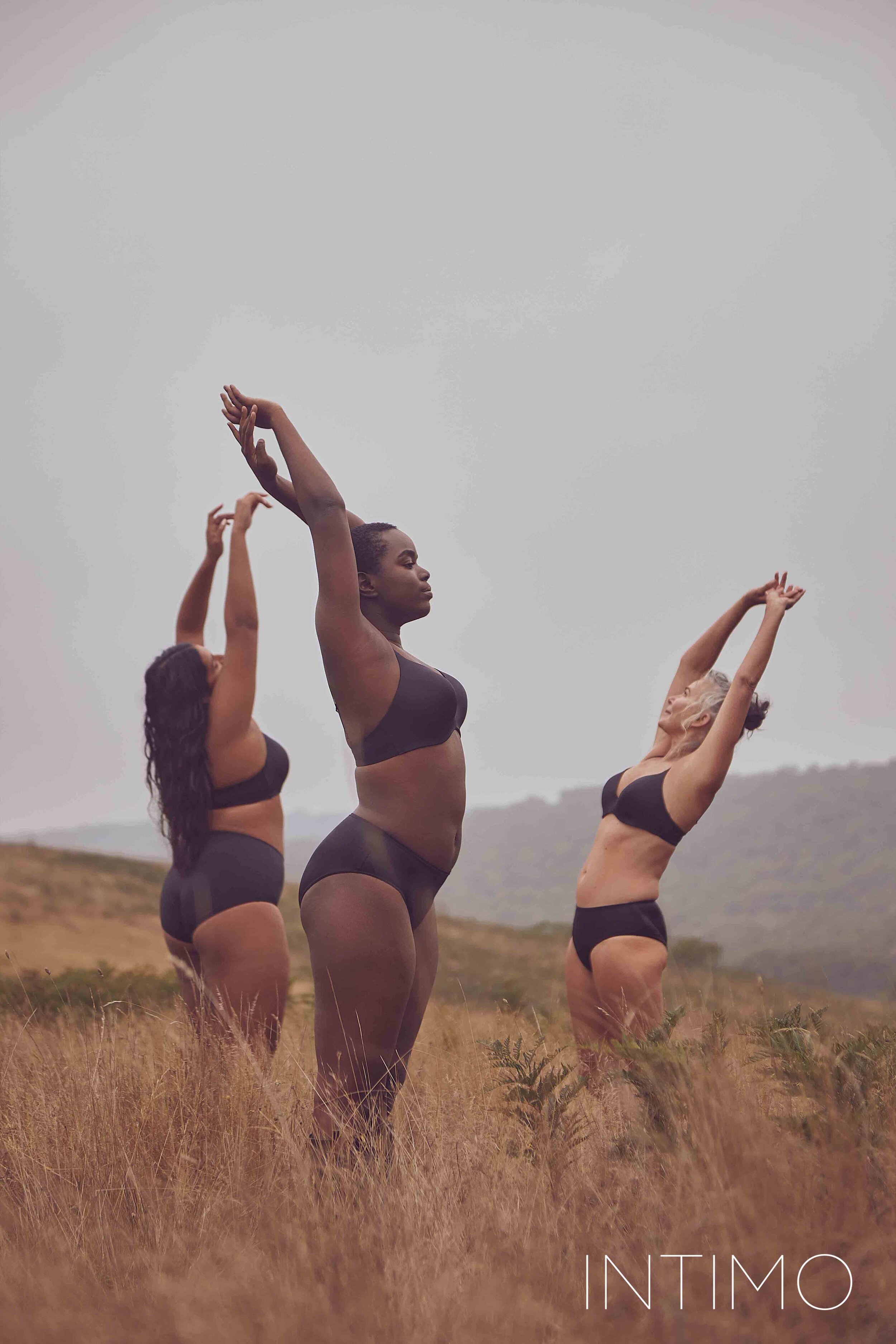 Three women in black underwear standing with arms raised in a grassy field beneath an overcast sky in an outdoor fashion lifestyle setting.