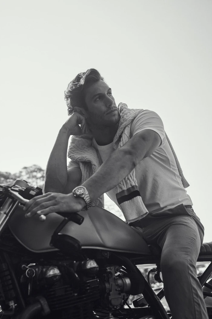 Monochrome portrait of a man leaning on a motorcycle wearing a t-shirt, trousers, and a sweater draped over his shoulders.