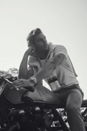 Monochrome portrait of a man leaning on a motorcycle wearing a t-shirt, trousers, and a sweater draped over his shoulders.