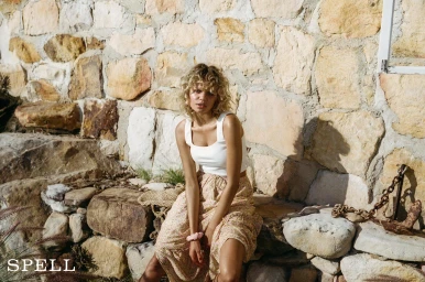 Blonde woman wearing a white tank top and floral skirt seated outdoors against a rustic stacked stone wall with an iron anchor prop.